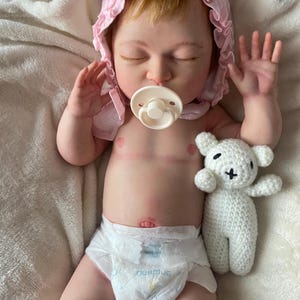 Puede incluir: Una mu&ntilde;eca beb&eacute; realista con los ojos cerrados, que lleva un gorro rosa y un pa&ntilde;al. La mu&ntilde;eca tiene un chupete en la boca y sostiene un peque&ntilde;o osito de peluche blanco de ganchillo.