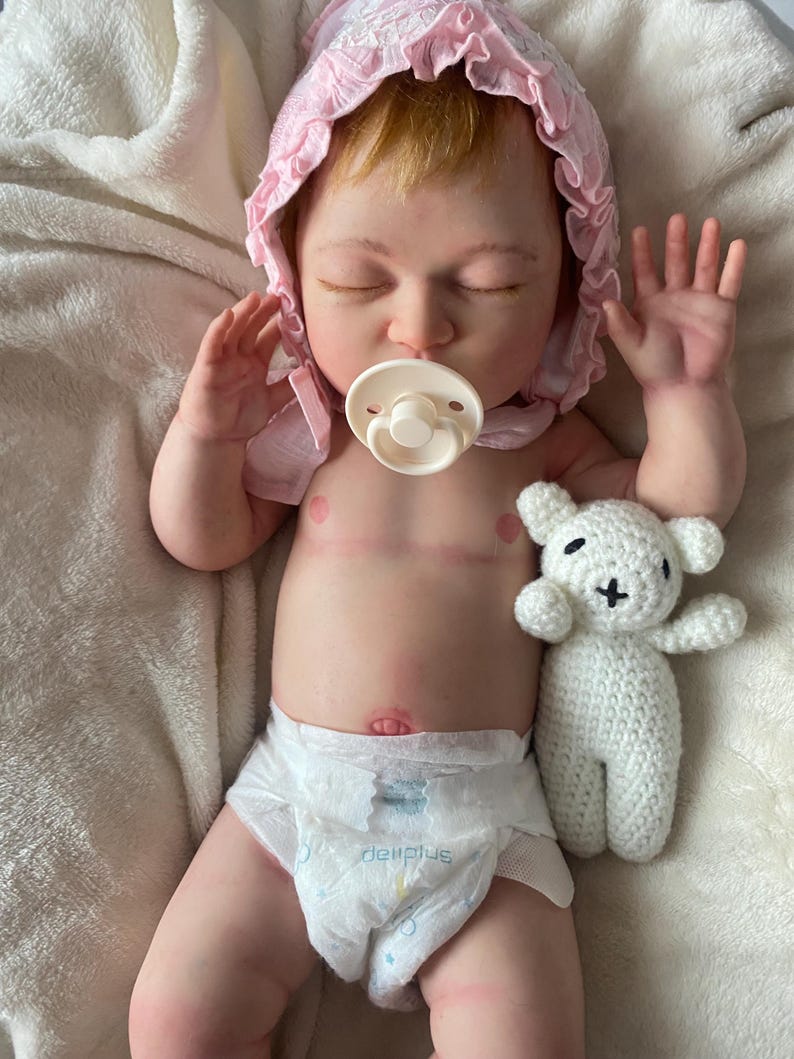 Puede incluir: Una mu&ntilde;eca beb&eacute; realista con los ojos cerrados, que lleva un gorro rosa y un pa&ntilde;al. La mu&ntilde;eca tiene un chupete en la boca y sostiene un peque&ntilde;o osito de peluche blanco de ganchillo.