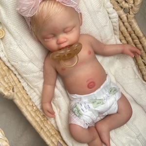 May include: A realistic newborn doll with light skin and blonde hair, wearing a pink lace bow headband and a white diaper. The doll is sleeping with a translucent pacifier in its mouth, resting on a white knitted blanket inside a woven basket.