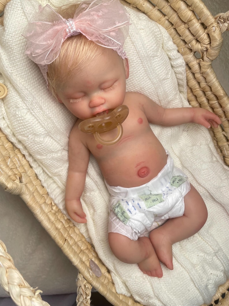 May include: A realistic newborn doll with light skin and blonde hair, wearing a pink lace bow headband and a white diaper. The doll is sleeping with a translucent pacifier in its mouth, resting on a white knitted blanket inside a woven basket.
