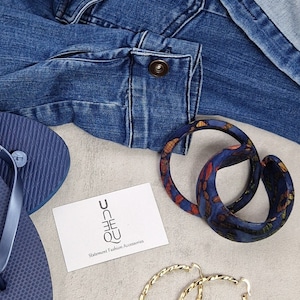 Könnte beinhalten: Zwei große, blaue und orangefarbene Ohrringe mit Muster, ein Paar goldene Ohrringe, ein blauer Flip-Flop, eine Jeansjacke und eine Visitenkarte mit dem Text "Unique Statement Fashion Accessories".