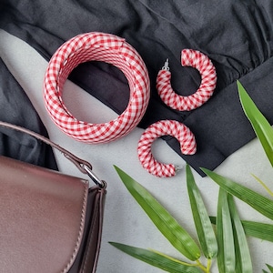 May include: Red and white gingham fabric earrings. The earrings are in the shape of a hoop and are approximately 5 cm in diameter. The earrings are displayed on a black fabric background.