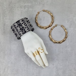 May include: A white mannequin hand wearing two gold rings and a black and white patterned bracelet. The hand is also wearing two large gold hoop earrings with a bamboo-like design.