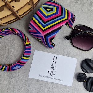 May include: Two colorful fabric bracelets with a geometric pattern in pink, blue, yellow, and black. The bracelets are on a gray surface with a white card that says "Un-Que Statement Fashion Accessories".