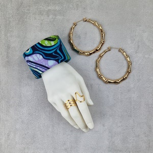 Puede incluir: Un maniquí de mano con dos anillos de oro y un brazalete de tela con estampado azul, verde y morado. El maniquí de mano también lleva dos grandes pendientes de aro dorados.