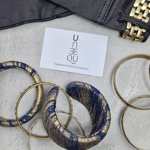 May include: A set of gold and blue patterned fabric bangles with a gold metal bangle. The bangles are arranged on a white surface with a business card that reads "UN-EQ Statement Fashion Accessories".