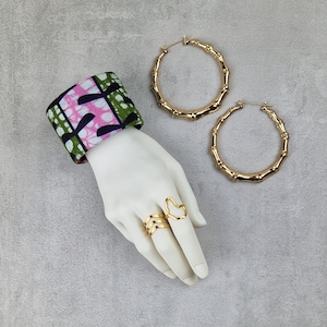 May include: A pair of gold bamboo hoop earrings and two gold rings on a mannequin hand wearing a pink and green patterned fabric wrist cuff.