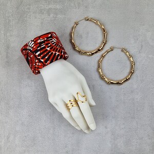 May include: A hand model wearing a red and black patterned fabric cuff bracelet and two gold rings. One ring is a simple band and the other is a stylized outline of the African continent. Two large gold hoop earrings with a bamboo-like design are also pictured.
