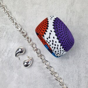 May include: A silver chain necklace with two silver teardrop-shaped earrings. The necklace is on a gray surface next to a colorful fabric bangle with a geometric pattern of red, white, blue, and black.