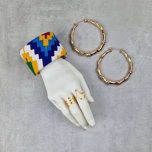 Può includere: Una mano di manichino bianca con due anelli dorati e un braccialetto in tessuto colorato. Gli anelli sono forme geometriche semplici. Il braccialetto ha un motivo geometrico in rosso, giallo, blu e bianco. La mano è su uno sfondo grigio.