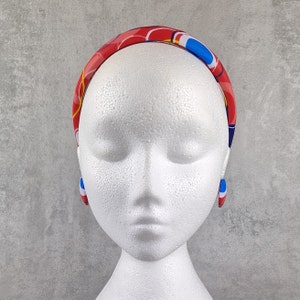 Op de afbeelding: Een witte mannequinkop met een rood, blauw en geel gestreept hoofdband met blauwe en witte oorbellen.