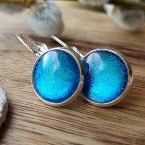 Könnte beinhalten: Ein Paar silberne Ohrhänger mit runden blauen Glascabochons. Die Cabochons haben eine glitzernde, irisierende Oberfläche.