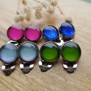 Könnte beinhalten: Eine Sammlung von acht runden Clip-Ohrringen mit glatten, gewölbten Oberflächen in verschiedenen Farben, darunter leuchtendes Pink, tiefes Blau mit Glitzer, schillerndes Hellblau und sattes Grün, alle in polierten, silberfarbenen Metallfassungen.