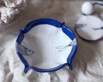 May include: A white ceramic dish with a blue rim and fish illustrations. The dish has a unique shape with curved edges and small feet. A matching round ornament with fish art is also present.