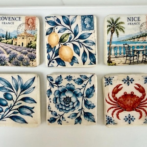 May include: Six square ceramic tiles featuring decorative illustrations. The top row shows a lavender field in Provence, lemons on branches, and a coastal scene in Nice. The bottom row displays olive branches, a blue floral pattern, and a red crab.