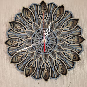 May include: A decorative wall clock with a mandala design. The clock has a layered, 3D effect and features a blue and brown colour scheme. The numbers 1 through 12 are displayed around the clock face.