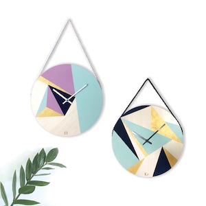 Peut inclure: Deux horloges murales rondes avec des motifs géométriques dans des tons de bleu, violet, or et noir. Les deux horloges ont une suspension en corde blanche.