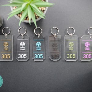 Puede incluir: Seis llaveros de acrílico transparente con anillas de metal plateadas. Cada llavero tiene un círculo blanco con el texto "YOUR LOGO" y el número "305" impreso en él. Los círculos son de diferentes colores: dorado, blanco, azul claro, negro, amarillo y rosa.