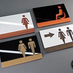 May include: Four acrylic signs with wood trim, each featuring a different restroom symbol. The signs are white with black symbols, or black with white symbols. The symbols include a woman, a man, a woman and a man together, and a person in a wheelchair. The signs are for a public restroom.
