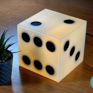 Può includere: Una lampada a forma di cubo progettata per assomigliare a un dado a sei facce con un corpo bianco traslucido e punti circolari neri. La lampada emette una luce soffusa dall'interno e presenta un cavo di alimentazione nero sul retro.