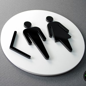 May include: White round sign with black silhouette figures of a man and woman. The sign has a black arrow pointing to the left.
