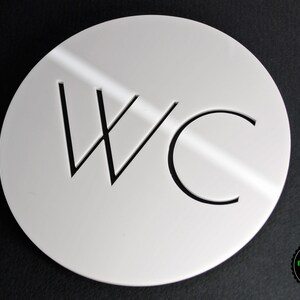 May include: White round sign with black lettering that says "WC" for a restroom.