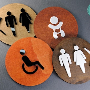 May include: Four round wooden signs with different symbols. The signs are brown, orange, and dark brown. The symbols are a man and woman with an arrow pointing left, a baby, a person in a wheelchair, and a man and woman with an arrow pointing right. The signs have the text "Premium Hotel Signs Quality" in a green circle.