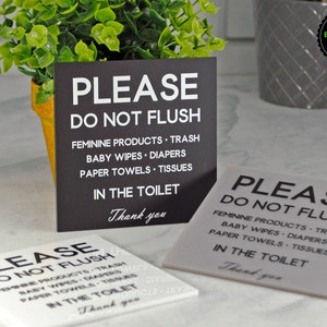 May include: Three white and black signs with the text "Please Do Not Flush" and a list of items that should not be flushed down the toilet. The signs are on a white surface.