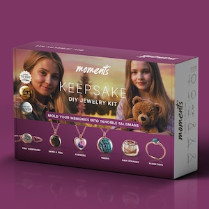 Op de afbeelding: Een doos voor een DIY-sieradenset genaamd "Moments Keepsake". De doos heeft een foto van twee meisjes, waarvan er één een teddybeer vasthoudt. De doos heeft een zilveren label met de tekst "Giet je herinneringen in tastbare talismannen". De doos toont zes verschillende sieraden, waaronder een ketting met een hartvormige hanger, een ketting met een ronde hanger, een ring en een hanger met een stoffen ontwerp. De tekst onder de sieraden luidt "Kleine aandenkens, Zand & aarde, Bloemen, Stof, Haarstrengen, Pluche speelgoed."
