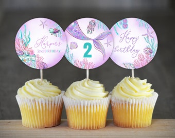 May include: Three purple and pink cupcake toppers with a mermaid theme. The toppers feature a mermaid tail, a starfish, and the text "Happy birthday". The toppers are on top of cupcakes with white frosting.