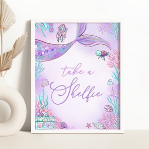Puede incluir: Cola de sirena morada y turquesa con un diseño de concha, estrella de mar y coral. El texto "take a shelfie" está escrito en cursiva morada.