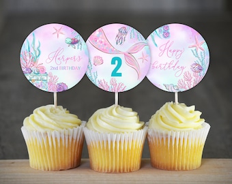 May include: Three cupcakes with pink and blue mermaid themed cupcake toppers. The toppers feature a mermaid tail, seashells, and the text "Happy birthday" and "Harper's 2nd Birthday".