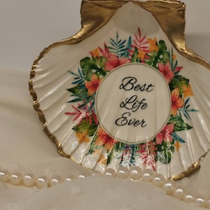 Könnte beinhalten: Eine weiße Muschel mit goldenem Rand und tropischem Blumendesign. Die Muschel trägt die Worte "Best Life Ever" in einem Kreis. Die Muschel liegt auf einer weißen Stoffoberfläche.