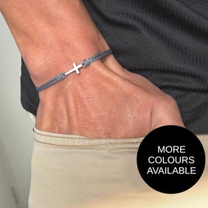 May include: A gray cord bracelet with a silver cross charm. The bracelet is worn on a wrist, with the text "MORE COLOURS AVAILABLE" in a black circle in the lower right corner.