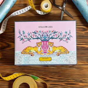 May include: A pink gift box with a blue border features a floral design with two tigers flanking a vase. The text "STILE D'CASA" and "GE LIVING" are printed on the box.