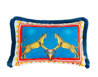 May include: A blue and orange patterned throw pillow with a fringed border features two leopards facing each other on a blue background. The leopards are flanking a gold vase.