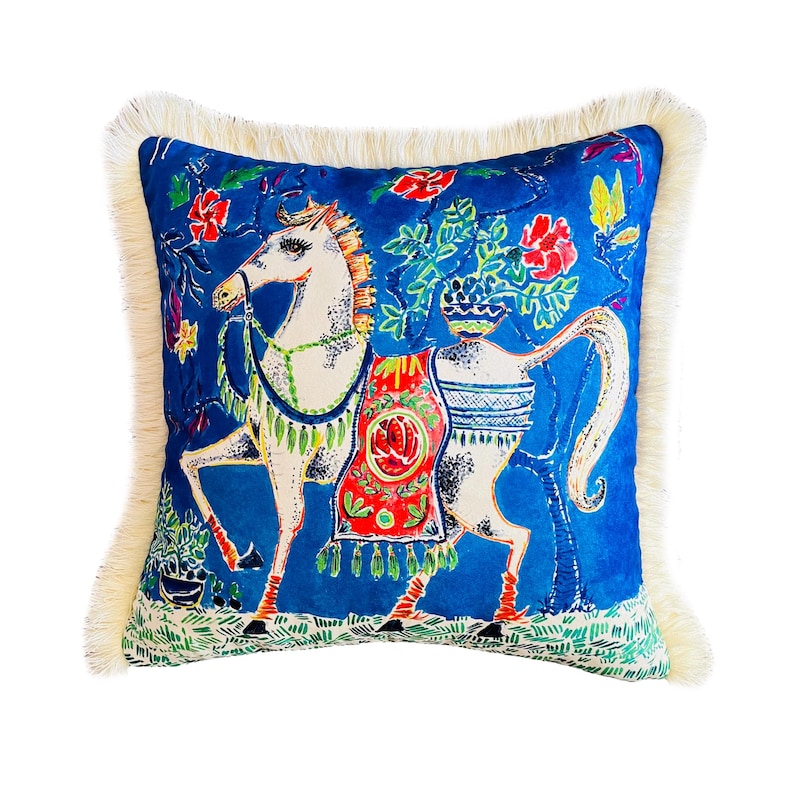 May include: A blue velvet pillow with a white fringe border features a colorful illustration of a white horse with a red, green, and yellow saddle. The horse is surrounded by green foliage and red flowers.