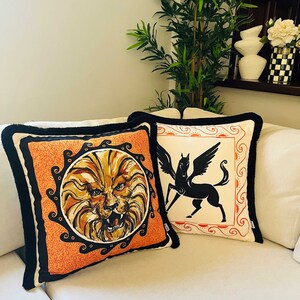 May include: Two decorative throw pillows with black trim. One pillow features a lion's face in a circle on an orange background. The other pillow features a black silhouette of a winged horse on a white background.