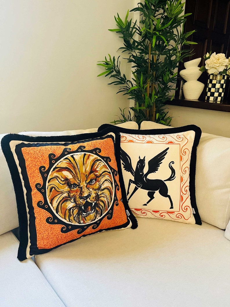 May include: Two decorative throw pillows with black trim. One pillow features a lion's face in a circle on an orange background. The other pillow features a black silhouette of a winged horse on a white background.