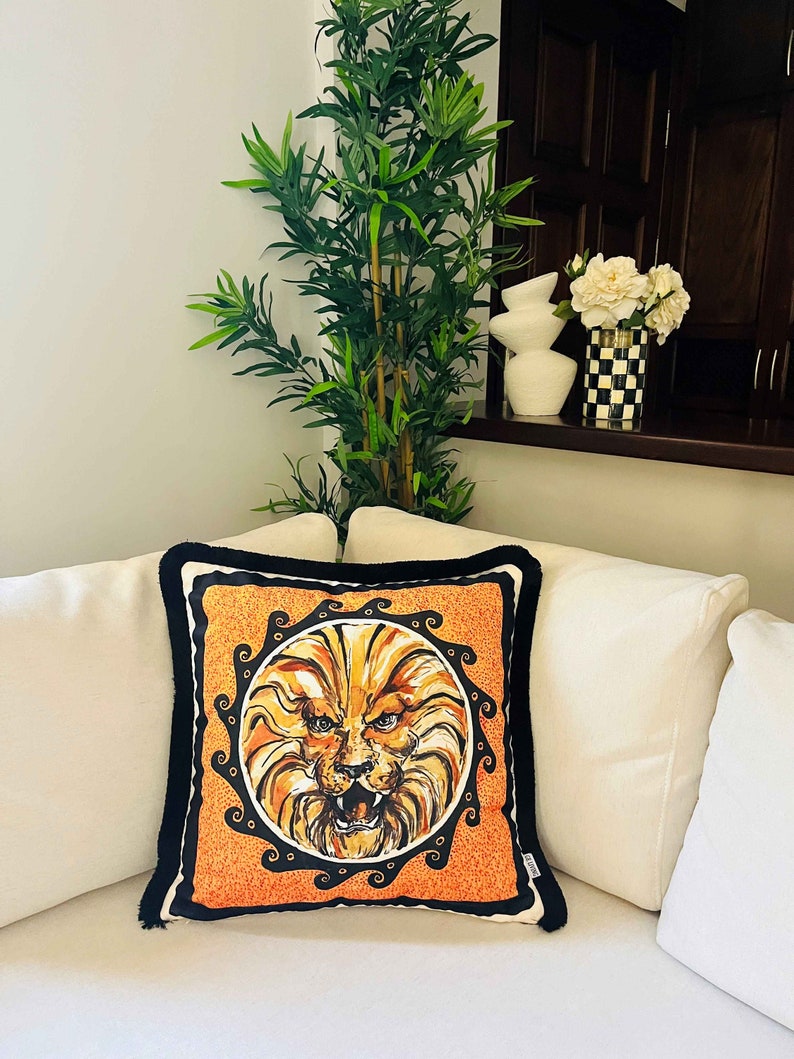 May include: A decorative throw pillow with a black trim features a lion's face in a circular frame with a black and orange background.