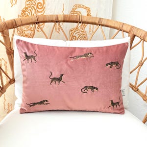 Puede incluir: Un cojín rectangular de terciopelo rosa con diseños de leopardo bordados. El cojín presenta múltiples figuras de leopardo en varias poses, sobre un fondo rosa suave. El cojín está colocado sobre un cojín blanco y una silla de mimbre.
