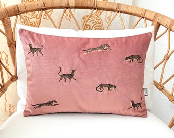 May include: A rectangular pink velvet pillow with embroidered leopard designs. The pillow features multiple leopard figures in various poses, set against a soft pink background. The pillow is placed on a white cushion and a wicker chair.