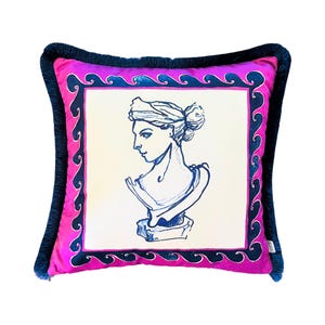 Op de afbeelding: Een roze kussen met een zwarte franje rand. Het kussen heeft een witte achtergrond met een blauwe en zwarte golfrand en een blauwe illustratie van een vrouwenbuste.