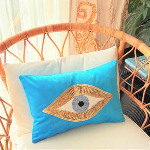 May include: A turquoise decorative pillow with a large eye design. The eye features a gold-colored, sequined outline, a light blue iris, and a black pupil. The pillow is placed on a white pillow and a wicker chair.