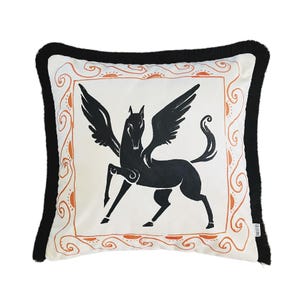Puede incluir: Una almohada blanca con un diseño de caballo alado negro dentro de un marco naranja y blanco. La almohada tiene un borde de flecos negro.
