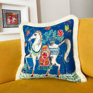 May include: A blue throw pillow with a white fringe border features a colorful illustration of a horse with a floral design. The horse is white with red, green, and yellow accents.