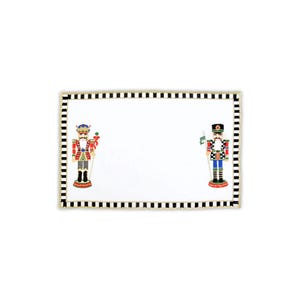May include: A white rectangular placemat with a black and white checkered border and gold trim. Two nutcracker figures are featured on the placemat, one on each side. The nutcracker on the left is wearing a red and white uniform with a blue and white hat. The nutcracker on the right is wearing a blue and white uniform with a red and white hat.
