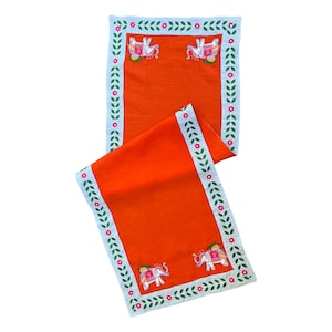 May include: Orange table runner with a white and green border featuring embroidered elephants. The border has pink flowers.