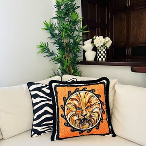 May include: Two decorative throw pillows on a white couch. One pillow has a black and white zebra print. The other pillow is orange with a black and gold lion face design.