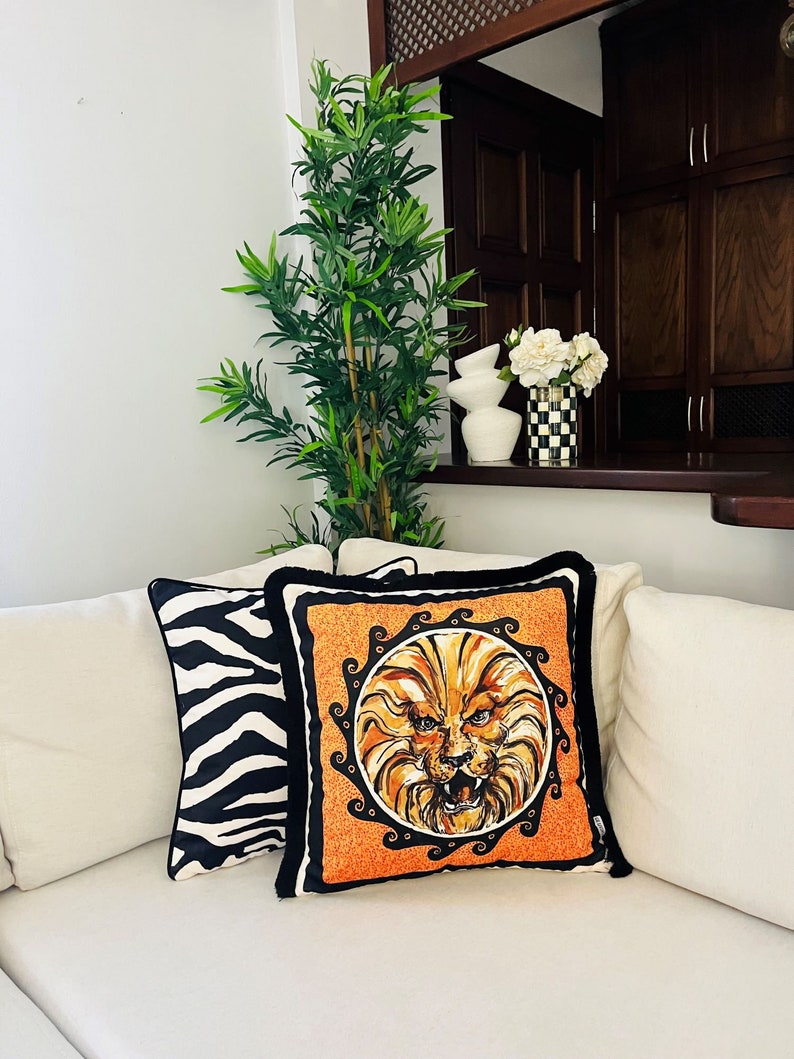May include: Two decorative throw pillows on a white couch. One pillow has a black and white zebra print. The other pillow is orange with a black and gold lion face design.